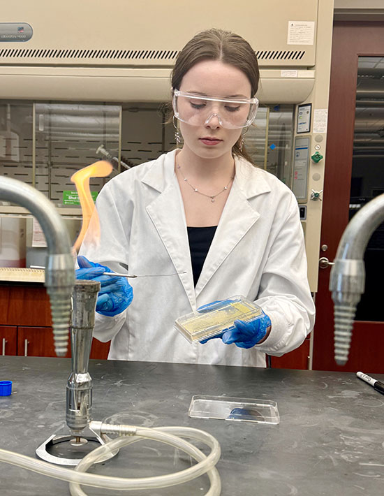Destiny M. O’Neill at a lab table with a lit bunson burner and lab equipment